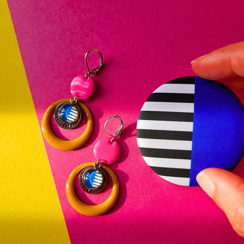 May include: A pair of earrings with yellow hoops, pink beads, and blue and white circular accents. A round, black and white striped and blue object is held in a hand. The background is a vibrant mix of pink and yellow.