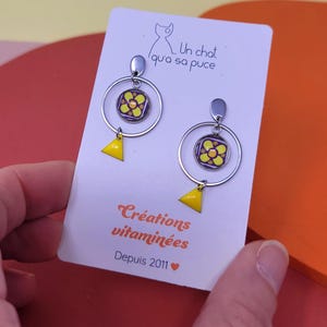 K&ouml;nnte beinhalten: Ein Paar silberne Creolen mit einem gelben Dreiecksanh&auml;nger und einem lila-gelben Blumenanh&auml;nger. Die Ohrringe befinden sich auf einer wei&szlig;en Karte mit dem Text "Un chat qui a sa puce" und "Cr&eacute;ations vitamin&eacute;es Depuis 2011".