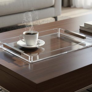 Pode incluir: Uma bandeja de servir de acrílico transparente com alças de aço inoxidável contém uma xícara e um pires brancos com uma colher. A xícara contém café escuro, e vapor sobe dela. A bandeja está em uma mesa de centro de madeira marrom escura.