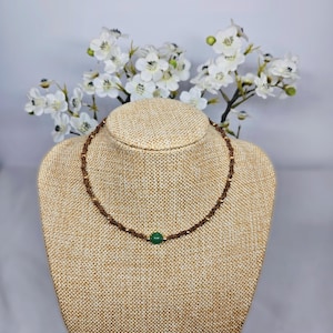 Peut inclure: Un collier de perles marron avec une pierre verte centrale. Le collier est présenté sur un buste à bijoux beige, avec des fleurs blanches en arrière-plan. Les perles sont petites et espacées uniformément.