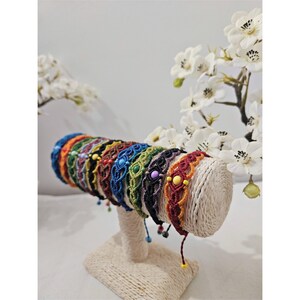 Peut inclure: Une exposition de bracelets en macramé colorés. Chaque bracelet présente des nœuds complexes dans diverses couleurs, notamment le bleu, le vert, le rouge et le noir, avec des perles décoratives. Les bracelets sont disposés sur un présentoir de couleur neutre, avec des fleurs blanches en arrière-plan.