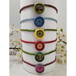 Könnte beinhalten: Eine Sammlung handgefertigter Makramee-Halsbänder in verschiedenen Farben, jedes mit einem einzigartigen zentralen Design. Die Halsbänder werden auf einem weißen zylindrischen Ständer präsentiert, der die komplizierten Details des Schmucks zeigt. Die Farben umfassen Braun, Blau, Grün, Rot und Lila.