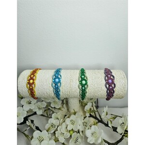 Op de afbeelding: Vier macramé armbanden in geel, blauw, groen en paars worden getoond op een witte, cilindrische standaard. De armbanden hebben een geweven ontwerp met kleurrijke kralen. Witte bloemen staan op de voorgrond.