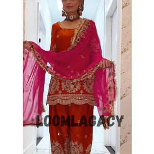 May include: A vibrant orange and red traditional outfit with gold embroidery. The ensemble features a long-sleeved top, matching trousers, and a sheer pink scarf. The text "LOOMLAGACY" is visible on the trousers.