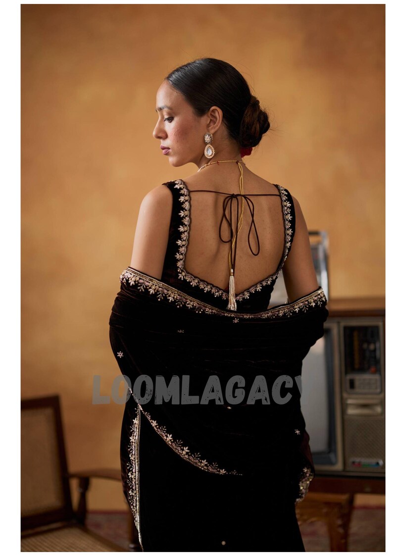 May include: A dark brown velvet sari with intricate gold embroidery. The sari features a low-cut back and a matching shawl. The text "LOOMLAGAC" is visible on the sari. The woman is wearing gold earrings and a necklace.