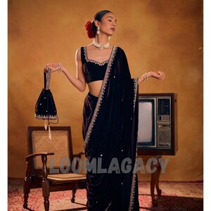 May include: A woman in a dark velvet saree with intricate embroidery and a matching velvet purse. The saree is draped elegantly, and the outfit is accessorized with jewelry and a red flower in the hair. The word "LOOMLAGACY" is visible on the image.