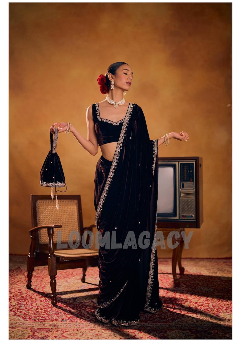 May include: A woman in a dark velvet saree with intricate embroidery and a matching velvet purse. The saree is draped elegantly, and the outfit is accessorized with jewelry and a red flower in the hair. The word "LOOMLAGACY" is visible on the image.