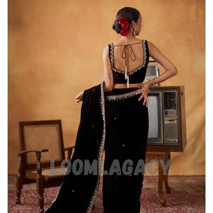 May include: A woman in a black velvet sari with intricate silver embroidery and embellishments. The sari features a matching black velvet blouse with delicate silver trim. Red flowers adorn the woman's hair, and the background has a vintage aesthetic.