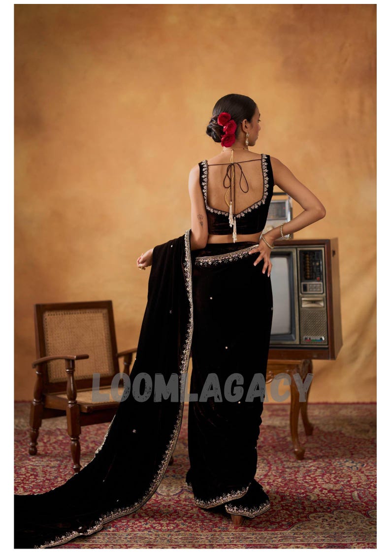 May include: A woman in a black velvet sari with intricate silver embroidery and embellishments. The sari features a matching black velvet blouse with delicate silver trim. Red flowers adorn the woman's hair, and the background has a vintage aesthetic.