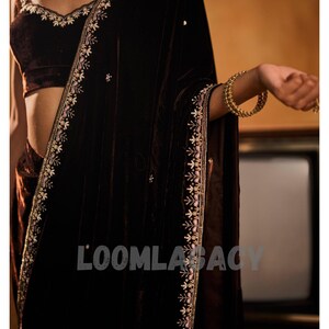 May include: A dark brown velvet saree with intricate embroidery along the edges and neckline. The saree features a matching velvet blouse with delicate floral patterns. Gold bangles adorn the wrist.