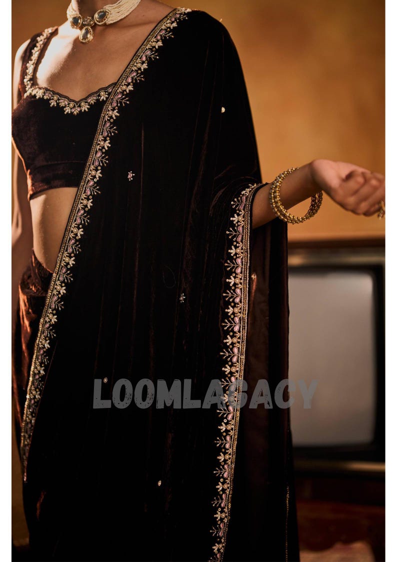May include: A dark brown velvet saree with intricate embroidery along the edges and neckline. The saree features a matching velvet blouse with delicate floral patterns. Gold bangles adorn the wrist.