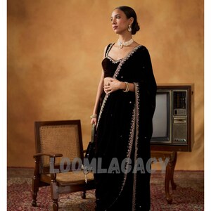 May include: A woman in a black velvet sari with intricate gold embroidery and embellishments. The sari is draped elegantly, paired with a matching velvet blouse. The background features a vintage television and a wooden chair.