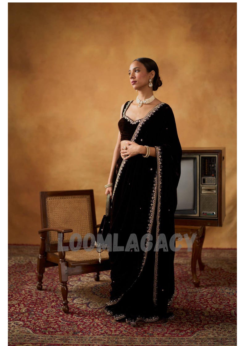 May include: A woman in a black velvet sari with intricate gold embroidery and embellishments. The sari is draped elegantly, paired with a matching velvet blouse. The background features a vintage television and a wooden chair.