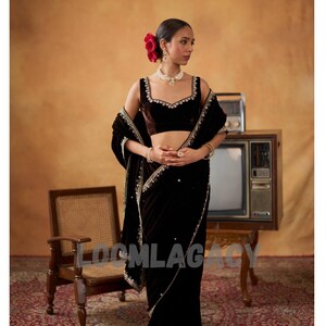 May include: A woman in a dark brown velvet saree with gold embroidery and a matching blouse. She wears a pearl necklace and earrings, with red flowers in her hair. The background includes a vintage TV and a wooden chair.