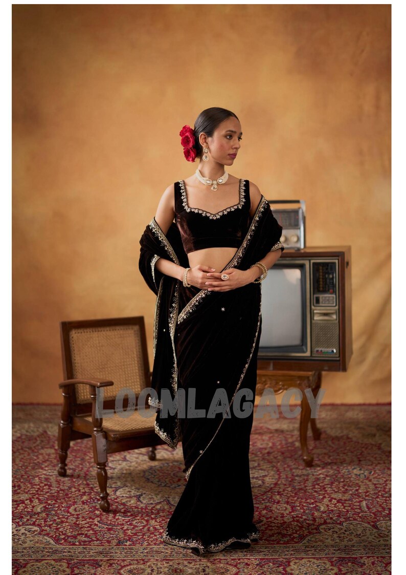 May include: A woman in a dark brown velvet saree with gold embroidery and a matching blouse. She wears a pearl necklace and earrings, with red flowers in her hair. The background includes a vintage TV and a wooden chair.