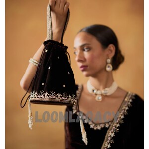 May include: A dark brown velvet drawstring bag with embroidered floral detailing in white and pink. The bag has a beaded handle and tassel accents. The word "LOOMLASACY" is printed on the bag.