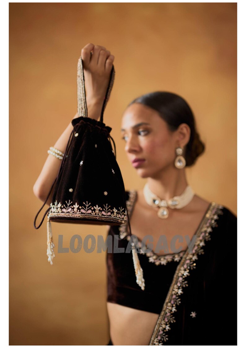 May include: A dark brown velvet drawstring bag with embroidered floral detailing in white and pink. The bag has a beaded handle and tassel accents. The word "LOOMLASACY" is printed on the bag.