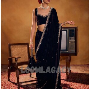 May include: A woman in a dark velvet sari with gold embroidery and a matching blouse. She is wearing jewelry and holding a small bag. A red flower adorns her hair. The background includes a vintage television and chair.