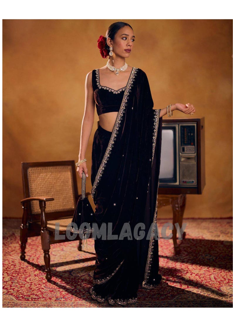 May include: A woman in a dark velvet sari with gold embroidery and a matching blouse. She is wearing jewelry and holding a small bag. A red flower adorns her hair. The background includes a vintage television and chair.