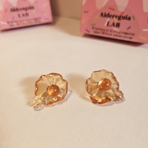 Puede incluir: Un par de pendientes con forma de flor, con pétalos transparentes de color ámbar y una cuenta central dorada y brillante. Los pendientes se muestran sobre una superficie blanca, con cajas rosas en el fondo.