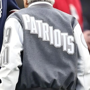 Könnte beinhalten: Grau-weiße Collegejacke mit dem Wort "PATRIOTS" in weißen Blockbuchstaben auf dem Rücken. Die Jacke hat weiße Ärmel, und die Zahlen "19" und "60" sind auf dem linken Ärmel zu sehen.
