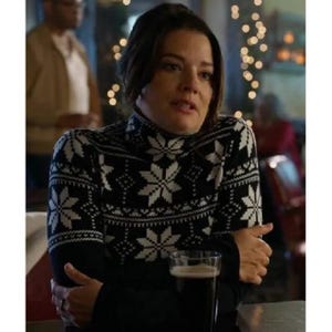 Könnte beinhalten: Ein schwarzer Rollkragenpullover mit weißem Schneeflockenmuster. Der Pullover hat lange Ärmel und einen hohen Ausschnitt. Ein Glas dunkles Bier ist im Vordergrund. Der Hintergrund ist verschwommen, mit einer Person und Lichtern sichtbar.