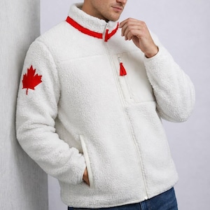 Puede incluir: Chaqueta de forro polar blanca con un emblema de hoja de arce roja en la manga izquierda. La chaqueta tiene un cuello alto con ribete rojo, una cremallera completa y un pequeño tirador de cremallera rojo. Tiene un bolsillo en el pecho y dos bolsillos laterales.