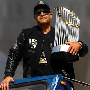 Könnte beinhalten: Eine Person trägt eine schwarze Jacke mit dem Text "Backtrack" und eine Baseballkappe und hält eine silberne und goldene Trophäe. Die Jacke hat Lederärmel und ein Logo auf der Brust. Die Trophäe hat einen silbernen Sockel und goldene Akzente.