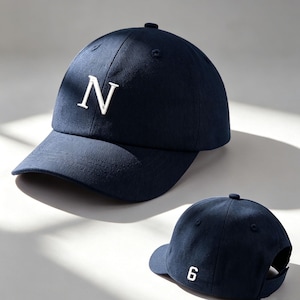 May include: A navy blue baseball cap featuring a white embroidered 'N' on the front. The rear of the cap shows the number '6' in white. The cap has a curved peak and a button on top.
