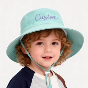 May include: A light teal sun hat with a wide brim and chin strap. The hat has the word "Custom" embroidered in purple script across the front. The hat is designed for children.