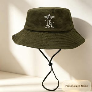 Puede incluir: Sombrero de pescador verde oliva con un diseño de faro blanco bordado. El sombrero tiene un ala ancha, una correa para la barbilla y un detalle de botón. El sombrero está hecho de una tela suave y texturizada.