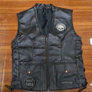Op de afbeelding: Een zwart leren vest met een ritssluiting aan de voorzijde, gevlochten afwerking langs de halslijn en veters aan de zijkanten. Het bevat een Harley-Davidson schedelpatch, een ronde patch met SINCE 1903 en een rechthoekige MOTORCYCLES-patch.
