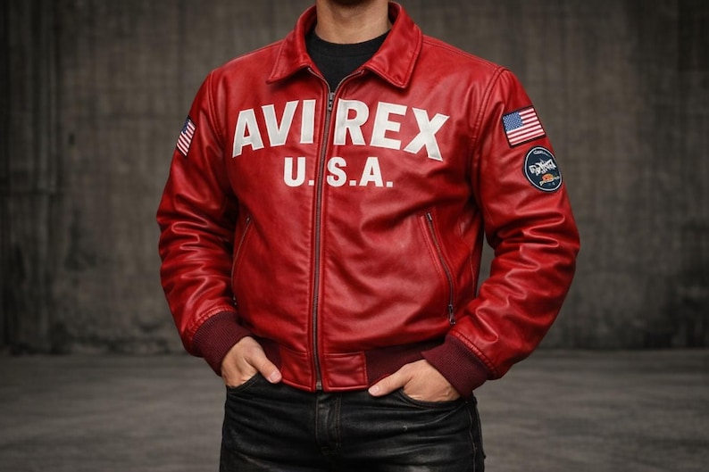 May include: A red leather jacket featuring a fold-down collar, a front zipper closure, and ribbed cuffs. The chest displays the white text AVI REX U.S.A. The sleeves feature an American flag patch and a circular logo patch on the left shoulder.