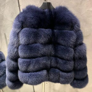 May include: A luxurious, navy blue fur jacket with a horizontal design. The jacket is made of soft, plush fur and has a round neckline. The jacket is hanging on a black hanger against a neutral background.