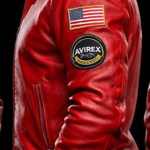 May include: A bright red leather jacket featuring a classic collar, a zippered side pocket, and a ribbed hem. The left sleeve displays a rectangular American flag patch positioned above a circular black and gold patch with the text AVIREX.