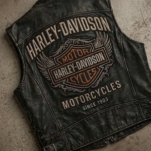 Könnte beinhalten: Eine schwarze Lederweste mit einem großen gestickten Harley-Davidson-Logo auf der Rückseite. Das Design enthält den Schriftzug Harley-Davidson, Motor Harley-Davidson Cycles, Motorcycles und Since 1903 sowie geflügelte Schildgrafiken in Beige und Orange.