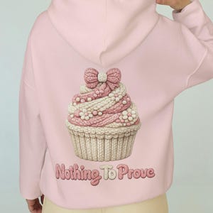 Peut inclure: Sweat à capuche rose clair avec un motif de cupcake détaillé dans le dos. Le cupcake présente un glaçage rose et blanc, un nœud et le texte "Nothing To Prove". Le sweat à capuche est de couleur rose clair unie.