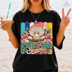 May include: A black t-shirt featuring a graphic print of a large open seashell containing a pearl with a gold crown. The design includes pink bows, turquoise text reading "PEARL GIRL" and "Summer" in white script, surrounded by pearls and starfish.