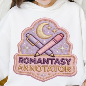 Könnte beinhalten: Weißer Kapuzenpullover mit einem gestickten Aufnäher. Der Aufnäher zeigt einen Halbmond, Sterne und gekreuzte Stifte. Der Text "ROMANTASY ANNOTATOR" ist in Lila und Gold gestickt, mit einem rosa und lavendelfarbenen Rand.