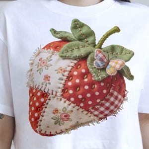 Puede incluir: Camiseta blanca con un gran diseño de fresa aplicado. La fresa es roja con lunares blancos y parches florales, hojas verdes y una pequeña mariposa. El diseño está cosido con costuras visibles.