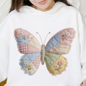 May include: White hoodie featuring a large, embroidered butterfly. The butterfly has patchwork wings in pastel colors, with floral and gingham patterns. The design includes embroidered flowers and a fringed edge.