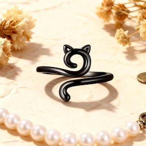 Puede incluir: Un anillo negro con forma de gato con un diseño de cola en espiral. El anillo presenta una cabeza de gato con orejas puntiagudas y una cola curvada que se envuelve alrededor del dedo. El anillo está sobre un fondo neutro con flores secas y un collar de perlas.