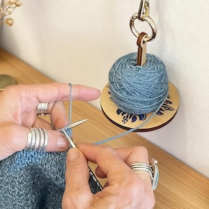 May include: A close-up of a knitting project in progress, featuring blue yarn and silver knitting needles. A wooden yarn holder with a blue ball of yarn hangs in the background. The yarn holder has a floral design.