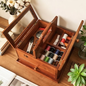 May include: A wooden sewing box with a glass lid, open to show spools of thread, scissors, and other sewing tools. The box is made of dark wood and has a drawer at the bottom. The box is on a wooden surface.