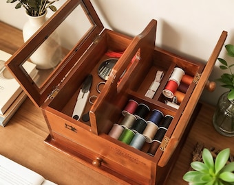 May include: A wooden sewing box with a glass-top lid, open to reveal spools of thread in various colors, scissors, and other sewing tools. The box is made of dark wood and has a drawer at the bottom.