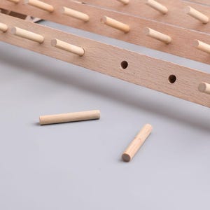 May include: Close-up of a wooden rack with several dowels. The rack is made of light-colored wood and has evenly spaced dowels protruding from it. Two loose dowels are lying on a gray surface. The rack appears to be a component of a larger structure.