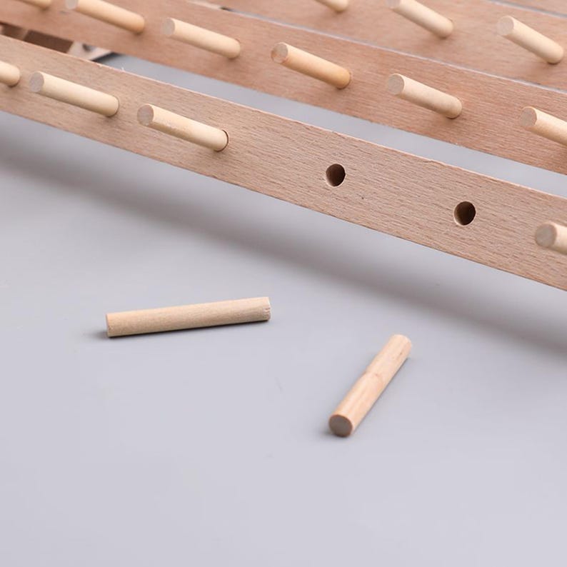 May include: Close-up of a wooden rack with several dowels. The rack is made of light-colored wood and has evenly spaced dowels protruding from it. Two loose dowels are lying on a gray surface. The rack appears to be a component of a larger structure.