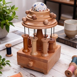 Pode incluir: Organizador de costura em madeira com almofada de alfinetes, suportes para bobinas e uma gaveta pequena. O organizador é feito de madeira castanha clara e apresenta um design em camadas com esferas de madeira e uma almofada de alfinetes com padrão floral.