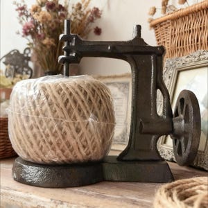 Può includere: Un distributore di spago in stile vintage in metallo scuro, simile a una vecchia macchina da cucire. Una grande palla di spago naturale è tenuta in posizione. Il distributore ha una ruota decorativa ed è posto su una superficie di legno.