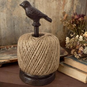 May include: A twine dispenser with a dark brown cast iron bird on top of a spool of natural twine. The twine is tightly wound, and the dispenser has a dark brown base. The background includes wooden panels and dried flowers.